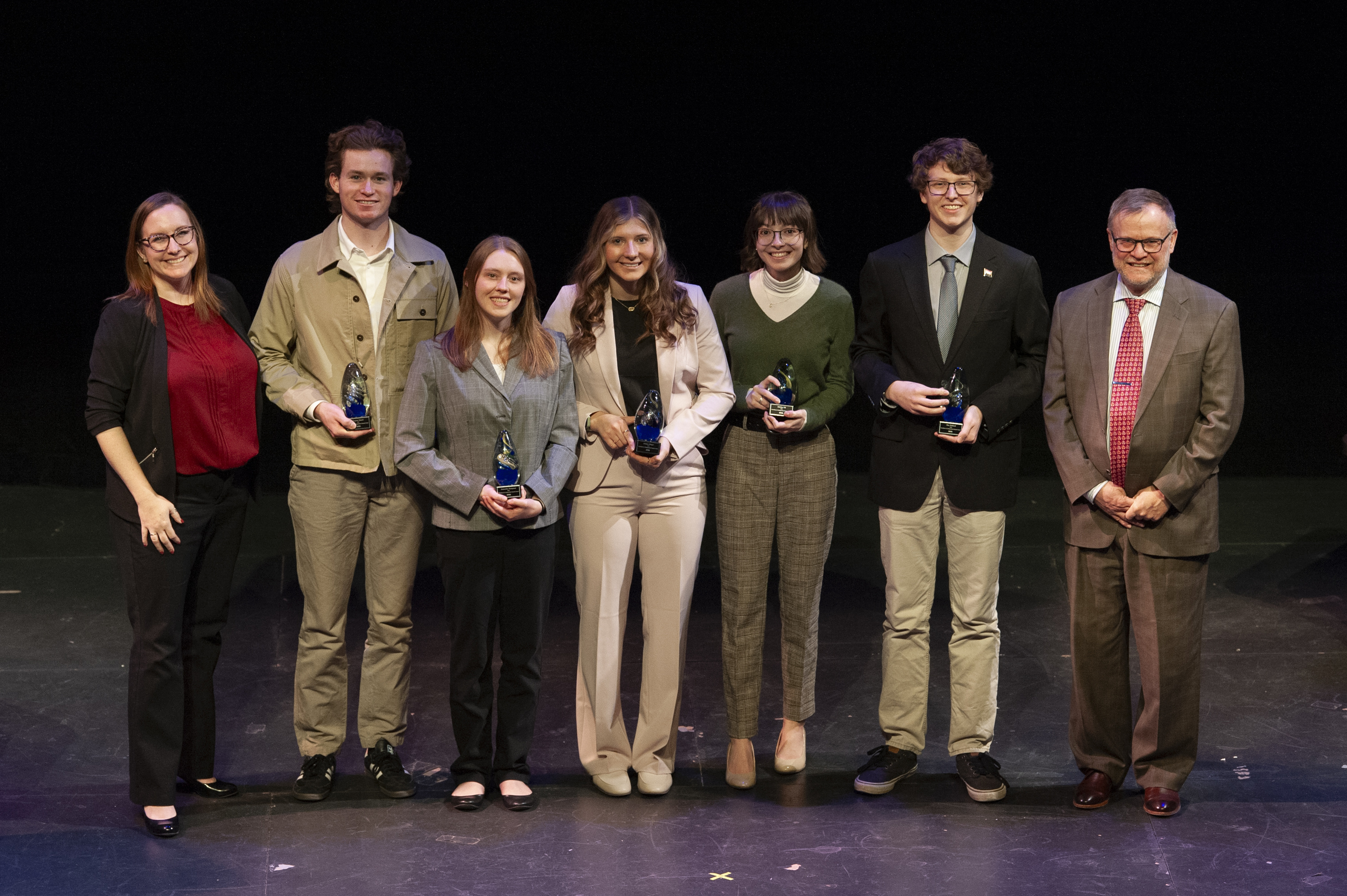 Five undergraduate students holding glass trophies on a theatre stage, flanked by Tawny Tibbits and Marty Scholtz.